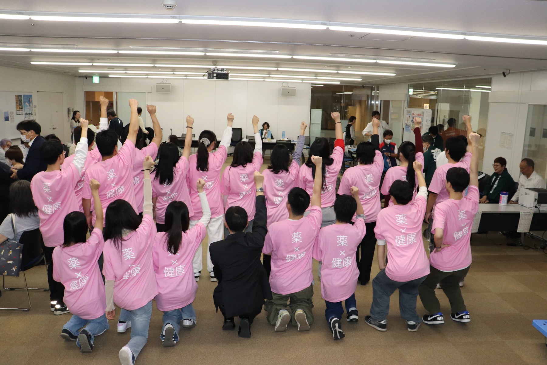 「薬×健康」と書かれたピンク色のTシャツを着て、背中を向けて片手を上げる学生たち